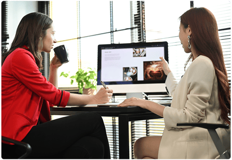 Two women are seated at a desk in an office, discussing content on a computer screen. One is holding a mug and taking notes, while the other gestures toward the screen. With natural light streaming through the window blinds, they focus on boosting efficiency through managed services.