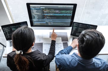 Two people are sitting at a table working on laptops, facing a large computer monitor displaying code. One person is pointing at the screen, highlighting their productivity and collaboration as an efficient IT team in a modern office setting.