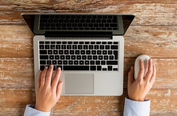 Top view of a person engaging with managed IT services on a laptop at a wooden table. One hand types on the keyboard, while the other holds a computer mouse. The laptop screen is open, reflecting an orderly workspace dedicated to tackling challenges efficiently.